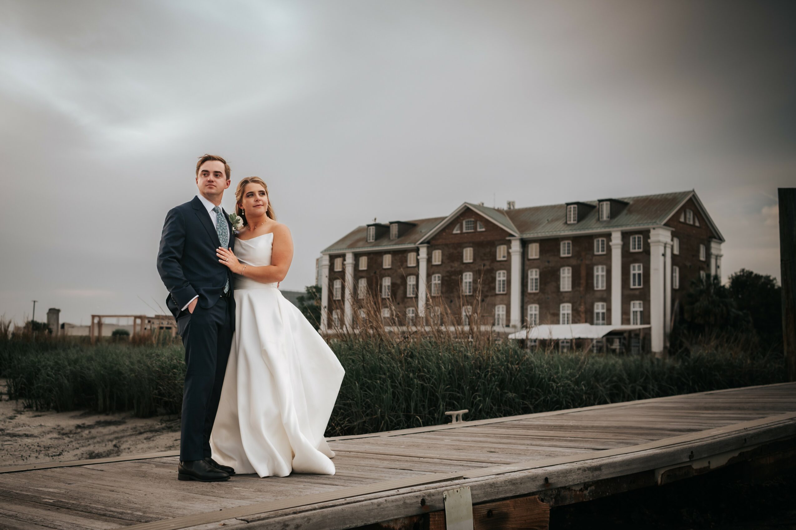 A couple in wedding attire standing on a wooden walkway outdoors with a large building in the background under cloudy sky.