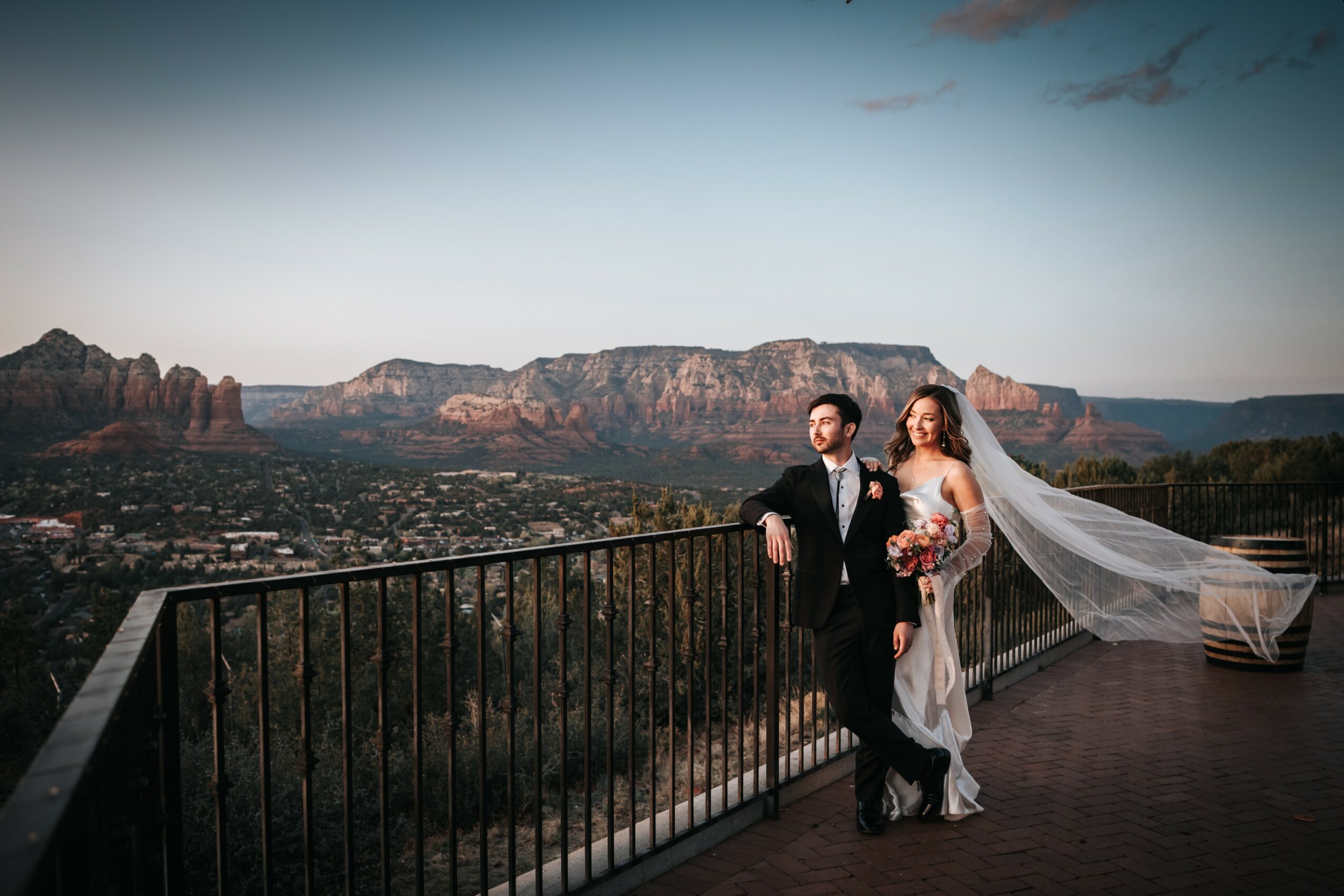 A bride and groom stand on a balcony with a mountain landscape in the background, holding a bouquet, during sunset.