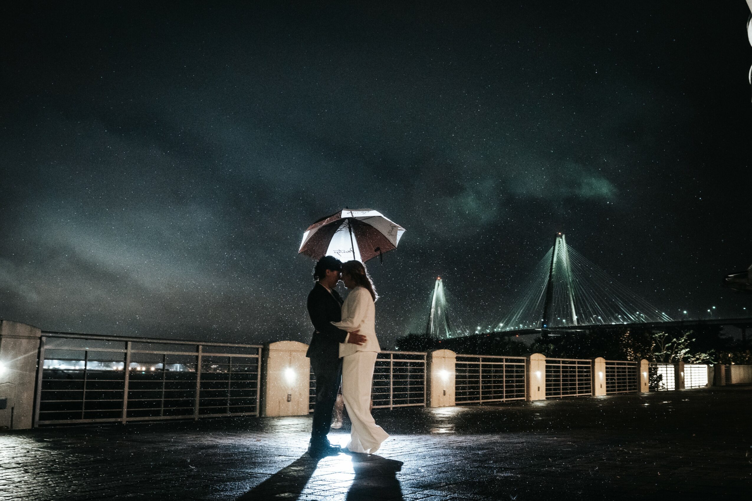 Two people stand close under an umbrella on a waterfront at night, with illuminated fountains and a cloudy sky in the background.