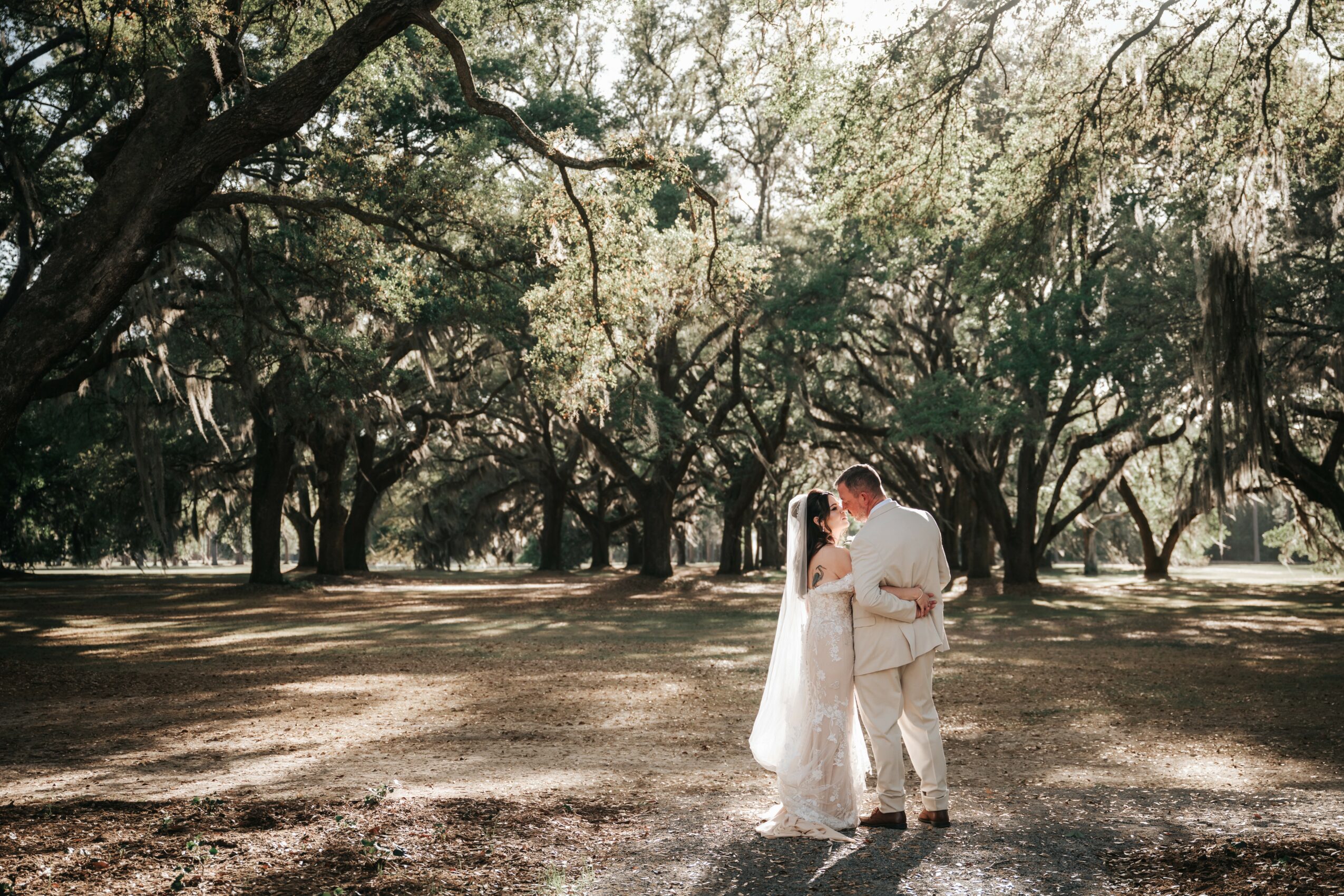 A couple dressed in wedding attire standing close together in a park with trees and sunlight filtering through the branches.