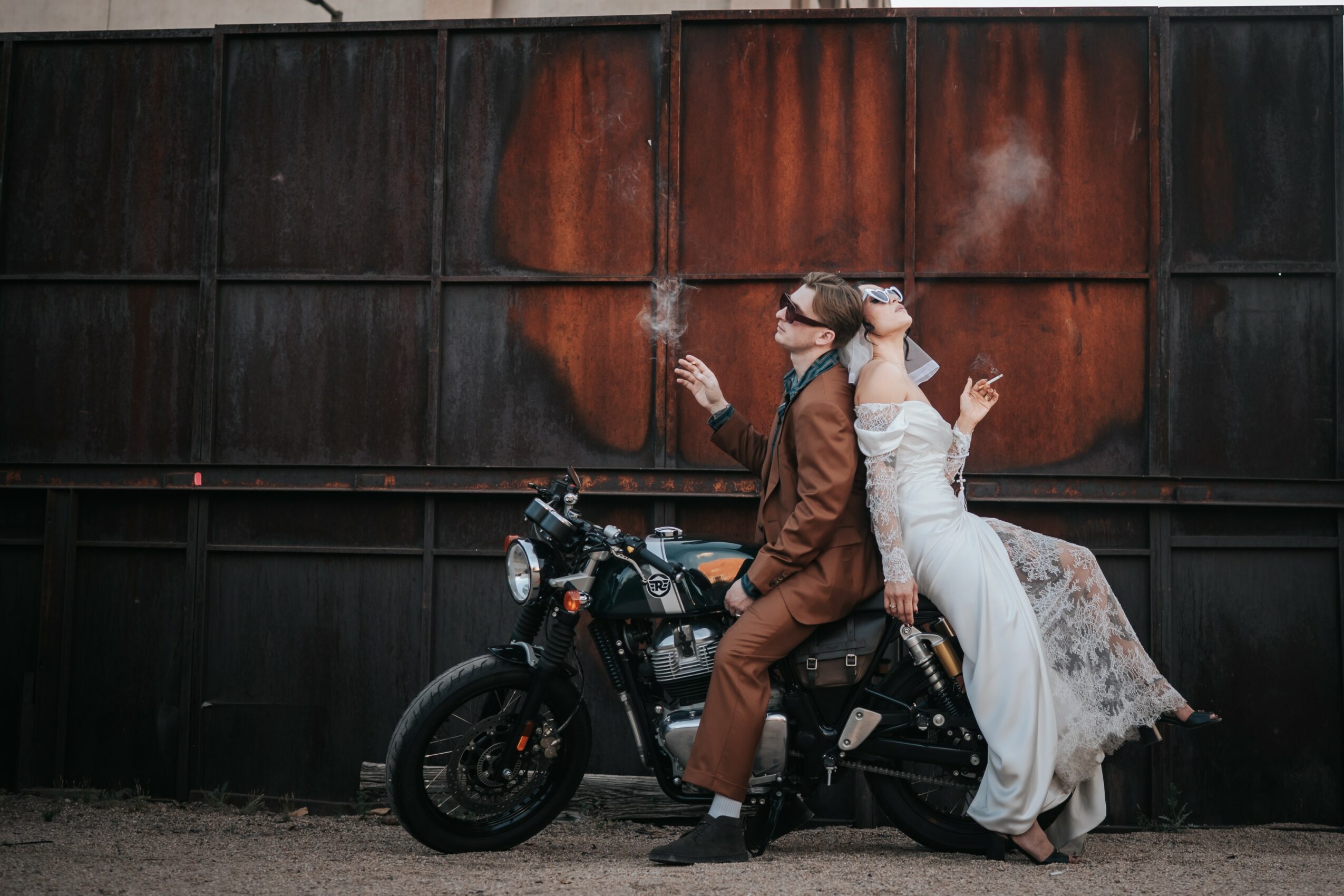 Two people sit back-to-back on a motorcycle in front of a large, rusty metal wall, one in a brown coat and the other in a white dress.