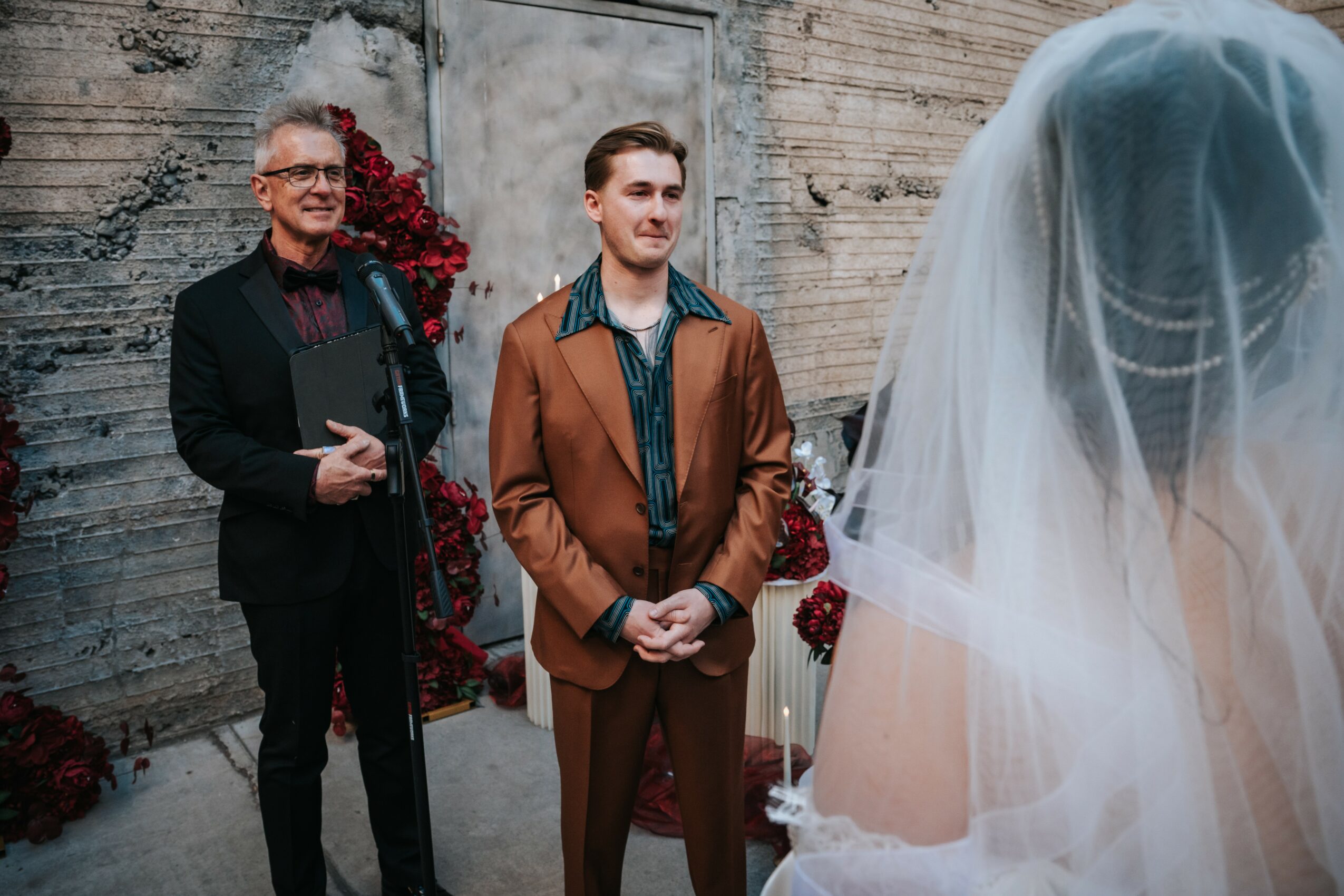 A man in a brown suit stands with hands clasped, facing a bride with a veil, in front of a textured wall.
