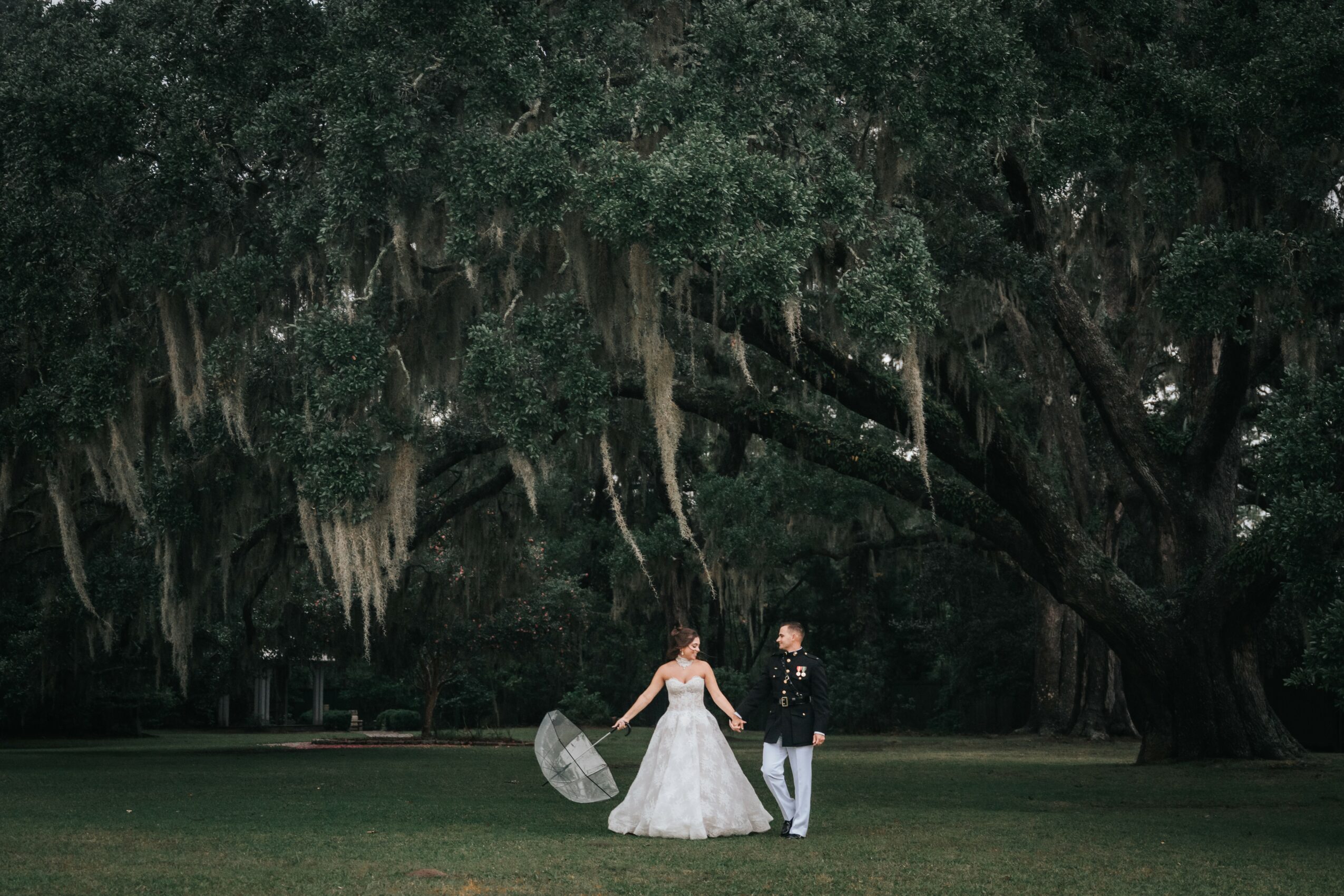 A couple holding hands in front of a large tree with hanging moss, one in a wedding dress and the other in formal attire.
