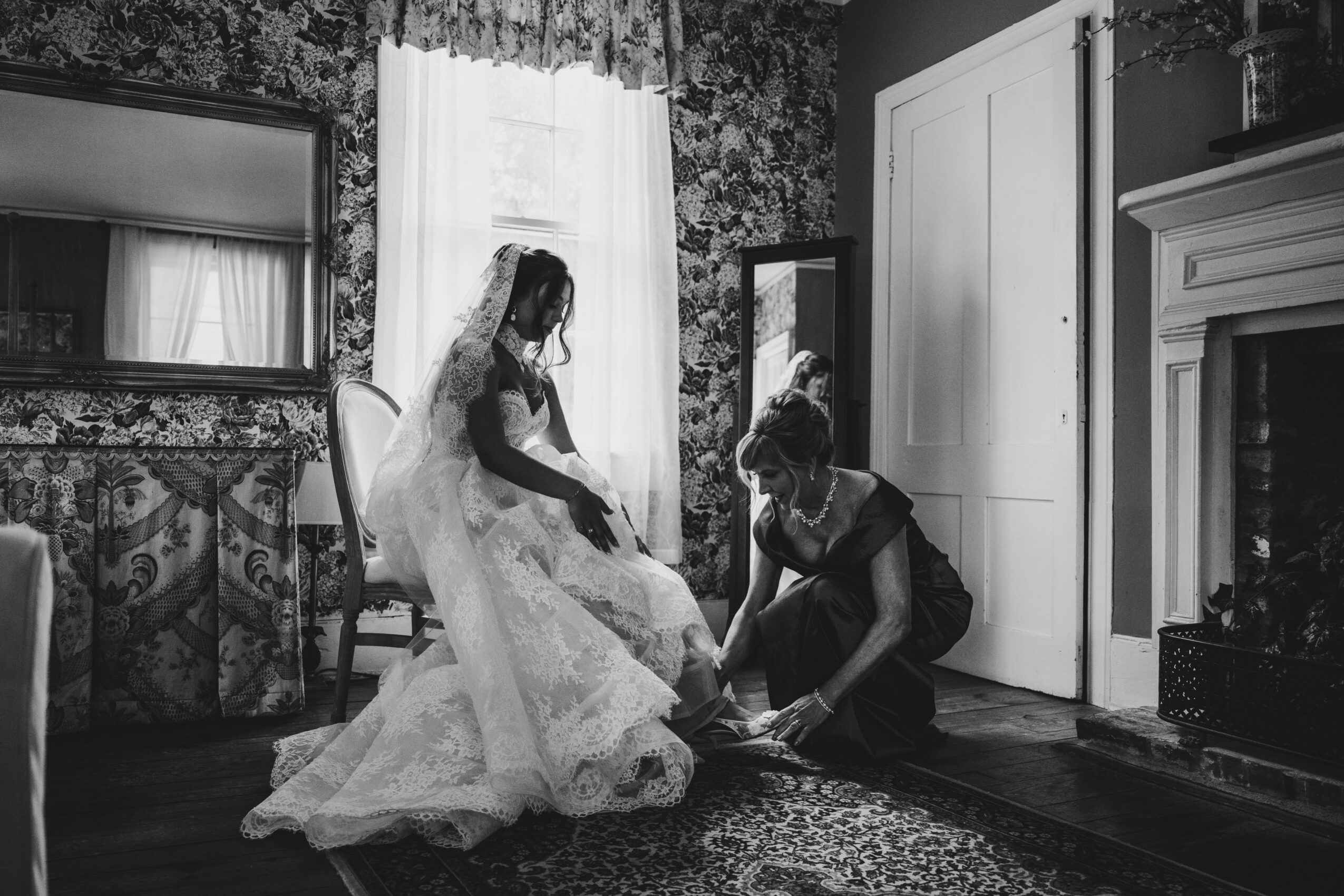 A bride in a wedding dress sits on a chair while a woman adjusts her shoe in a room with floral wallpaper and a fireplace.