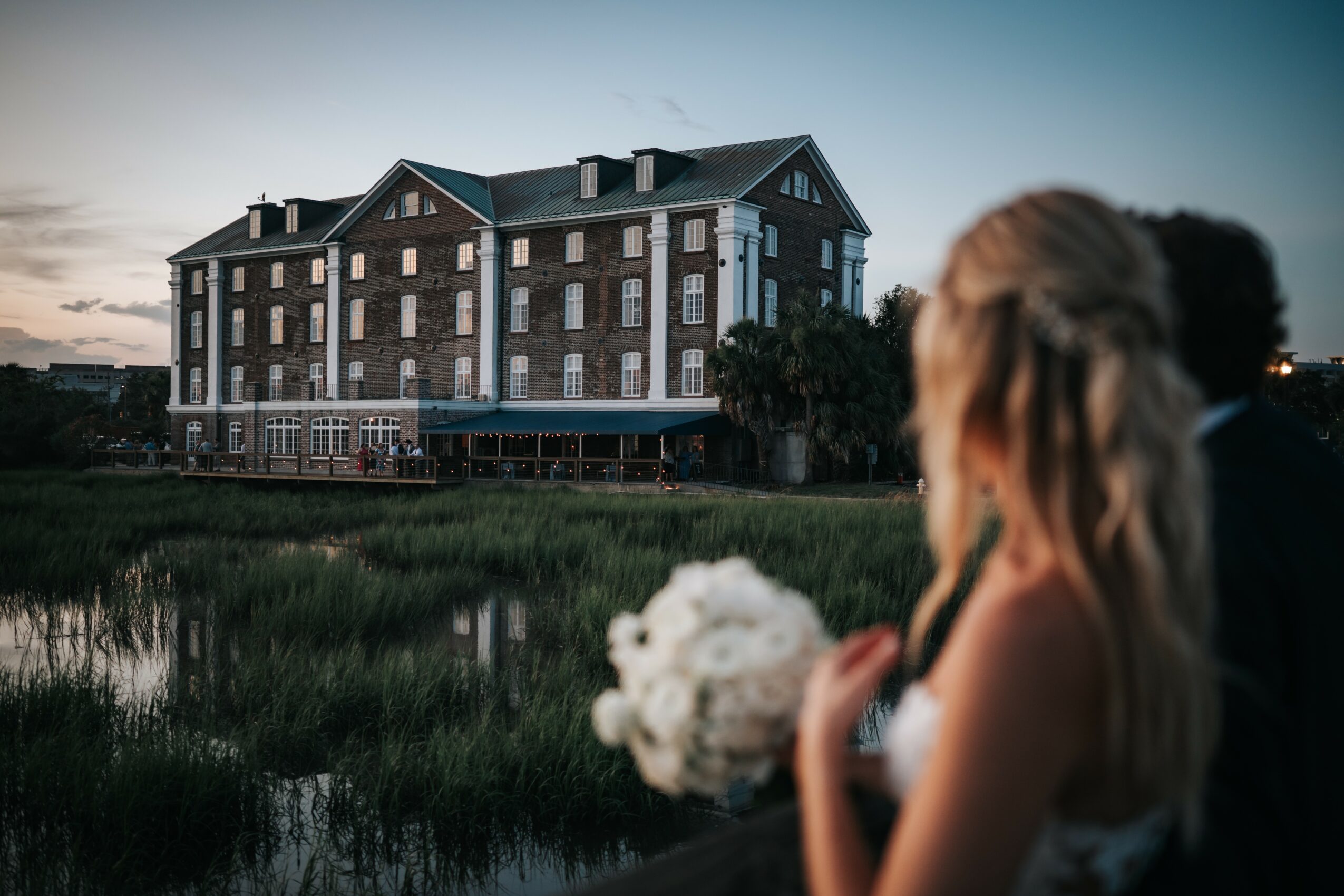 Person holding a bouquet of flowers outdoors near a large building with a river and greenery in the background.