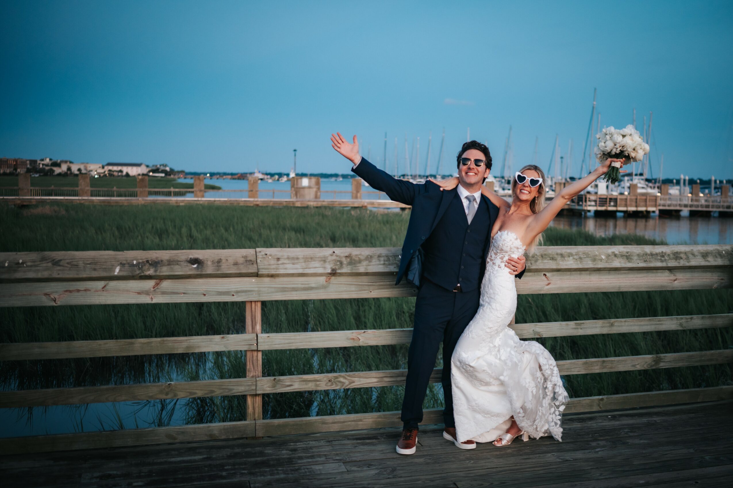 A bride and groom pose on a wooden dock with a marina and blue sky in the background, celebrating outdoors.