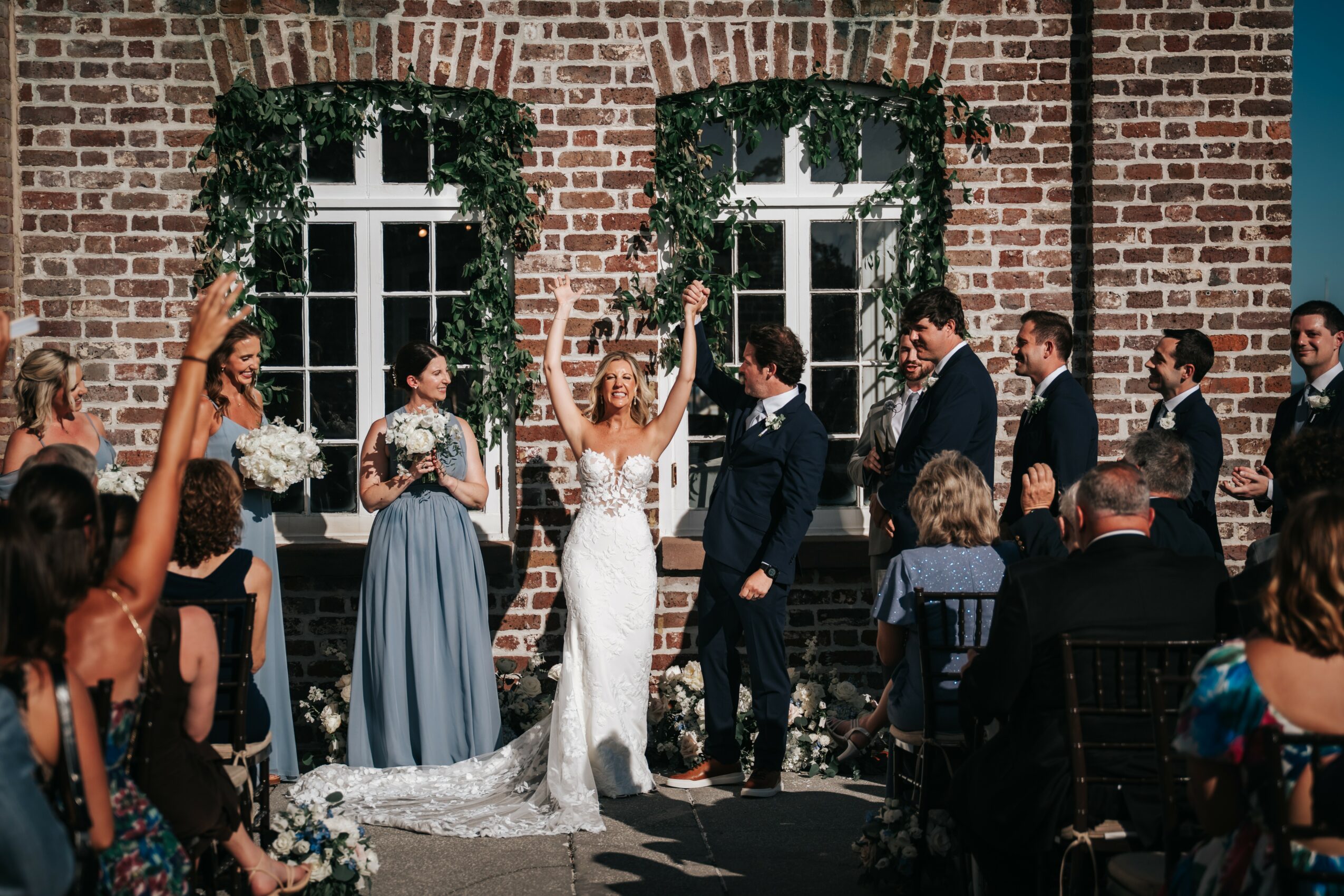 Bride and groom exchanging vows outdoors with wedding party and guests, brick building background, sunny day.
