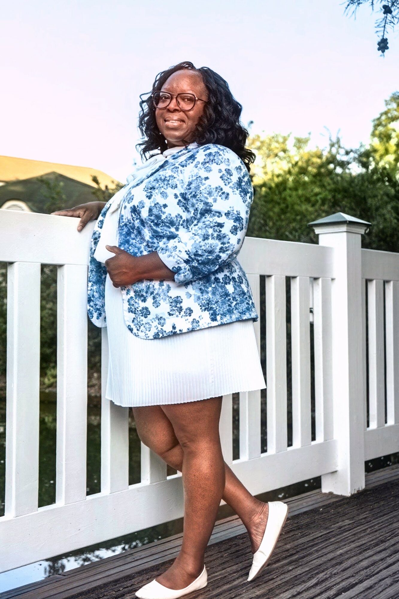 Woman standing outdoors near a white fence, smiling, wearing a blue floral jacket, white dress, and white shoes.