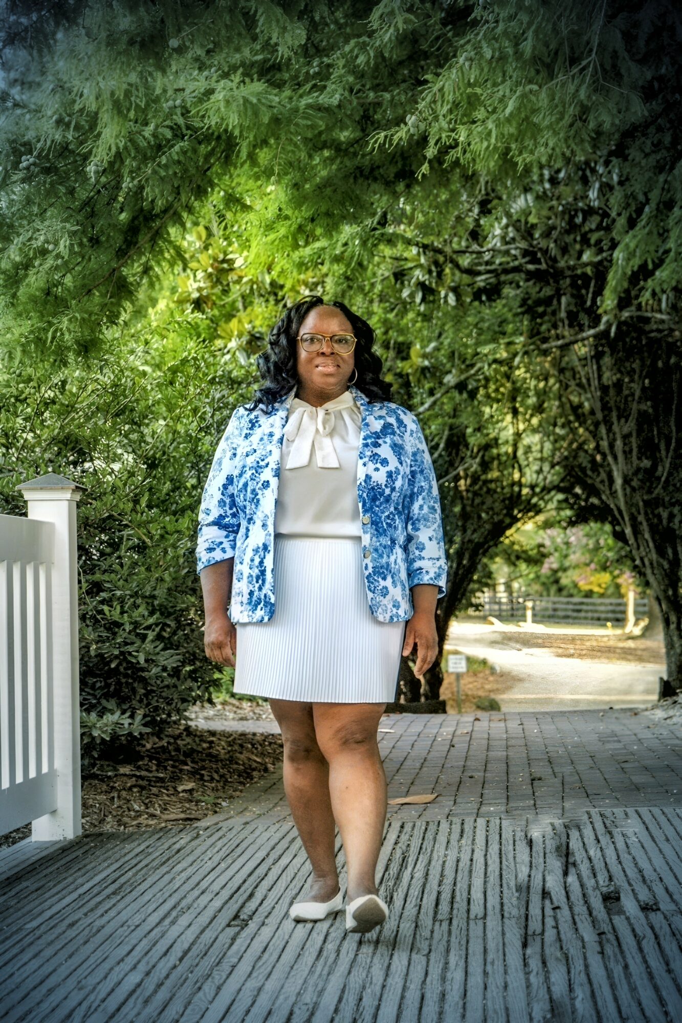 Woman standing outdoors on a paved path with trees in the background, wearing a white dress, blue patterned jacket, and white shoes.