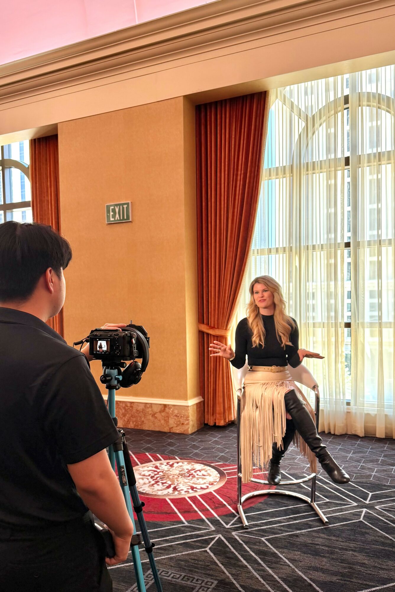 Woman sitting on a chair in a room with large windows and curtains, being filmed by a person with a camera.
