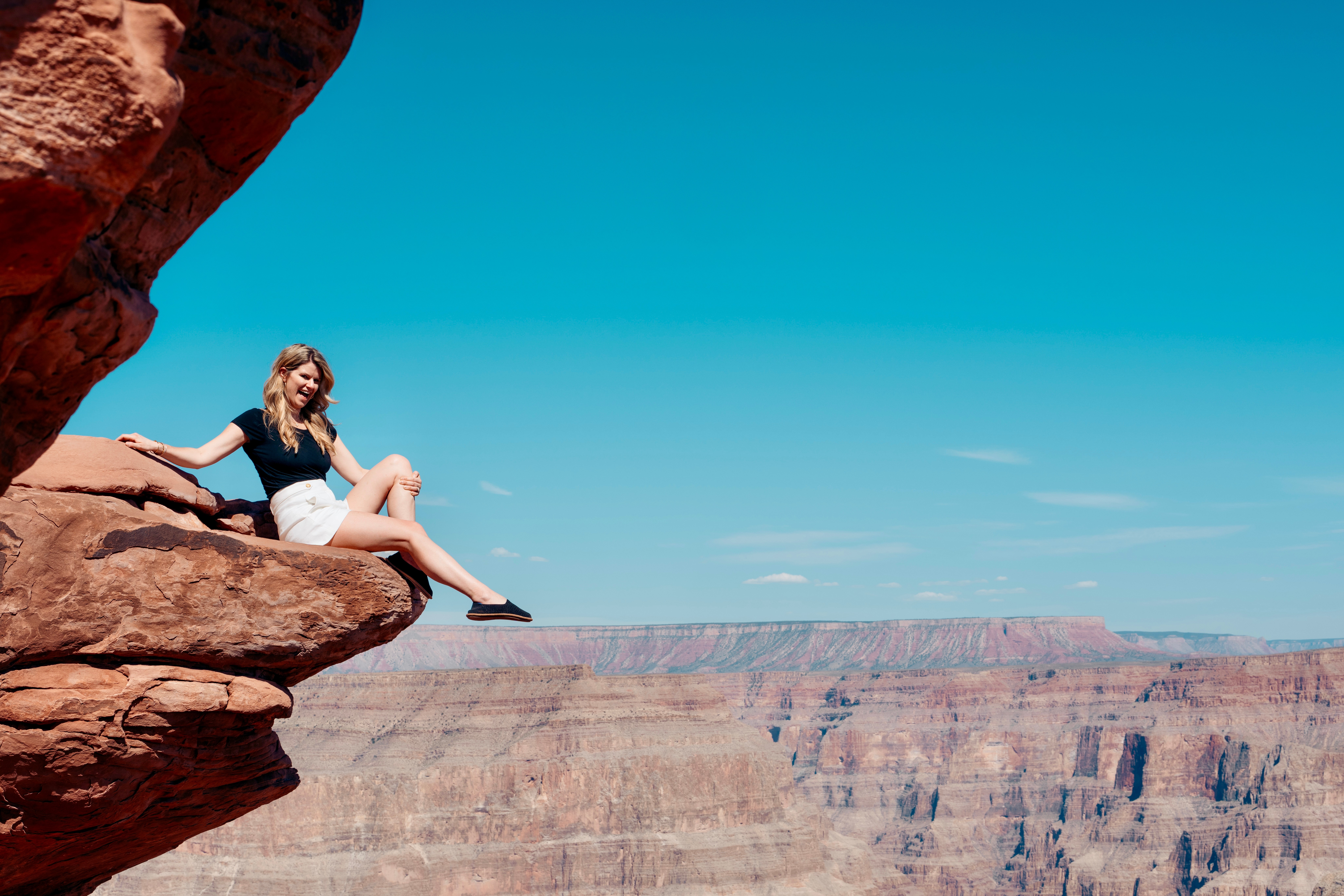 Person sitting on a rock ledge overlooking a canyon under a clear blue sky.