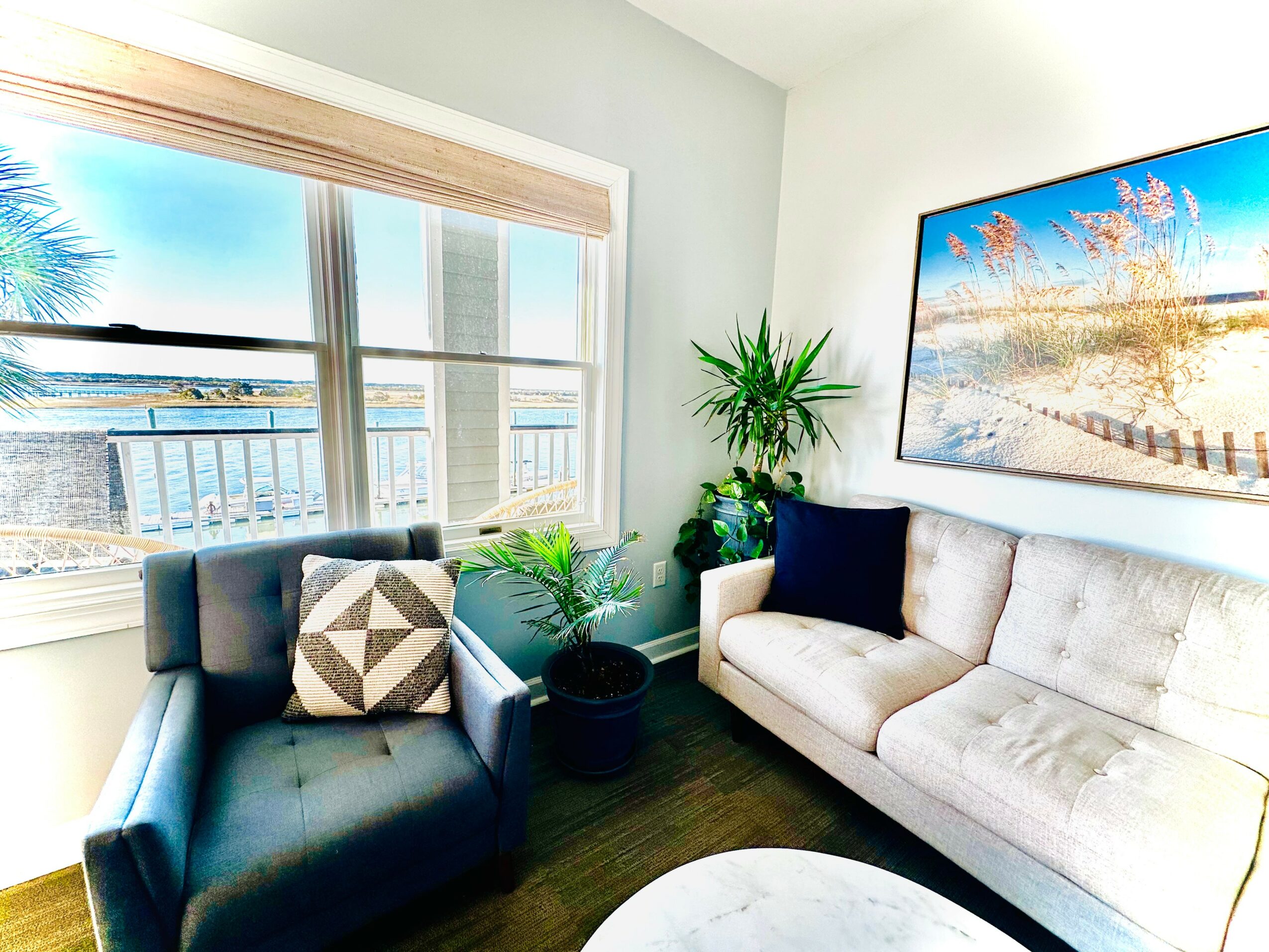 Living room with large window showing water and outdoor scenery, sofa, armchair, plants, and a wall-mounted TV.