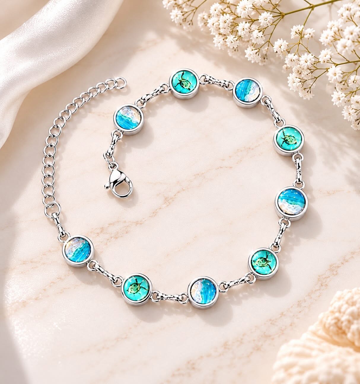 Silver bracelet with circular blue and green ocean-themed charms, laid on a marble surface with flowers nearby.