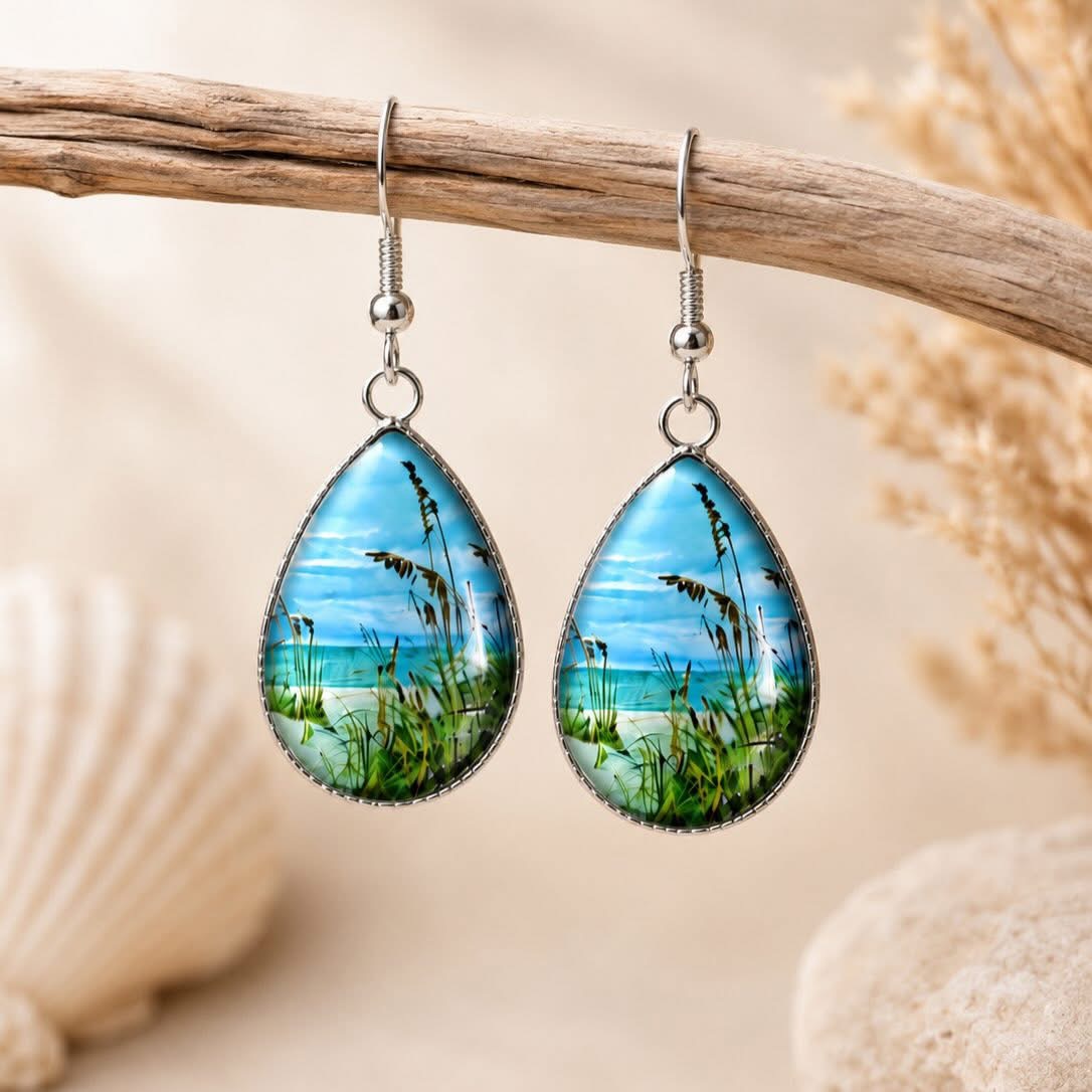 Pair of teardrop-shaped earrings with beach scene, grass, and sky, hanging from a wooden branch, neutral background.
