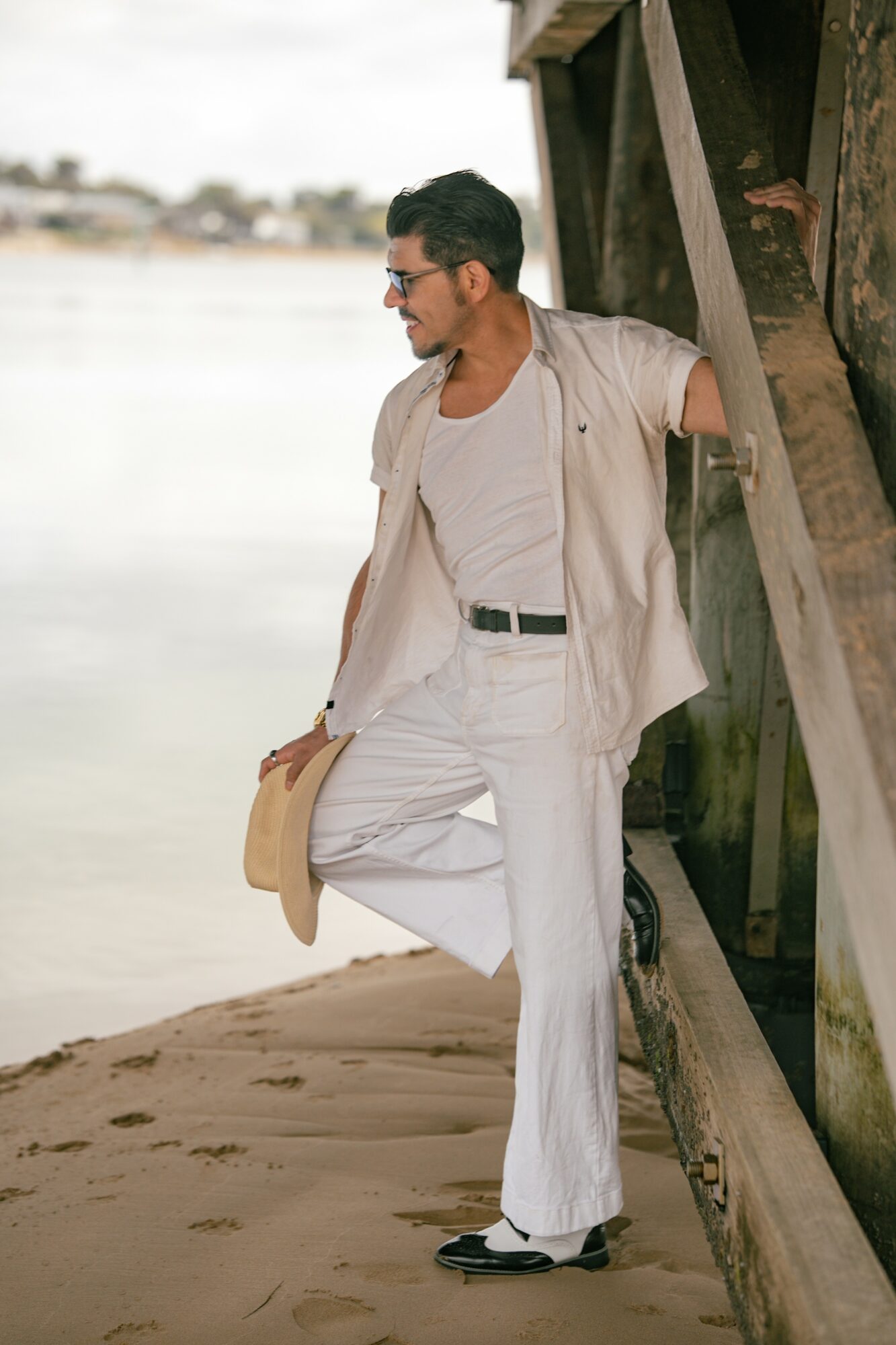 Man in white outfit leaning against a wooden structure on a sandy beach, holding a hat, with water in the background.