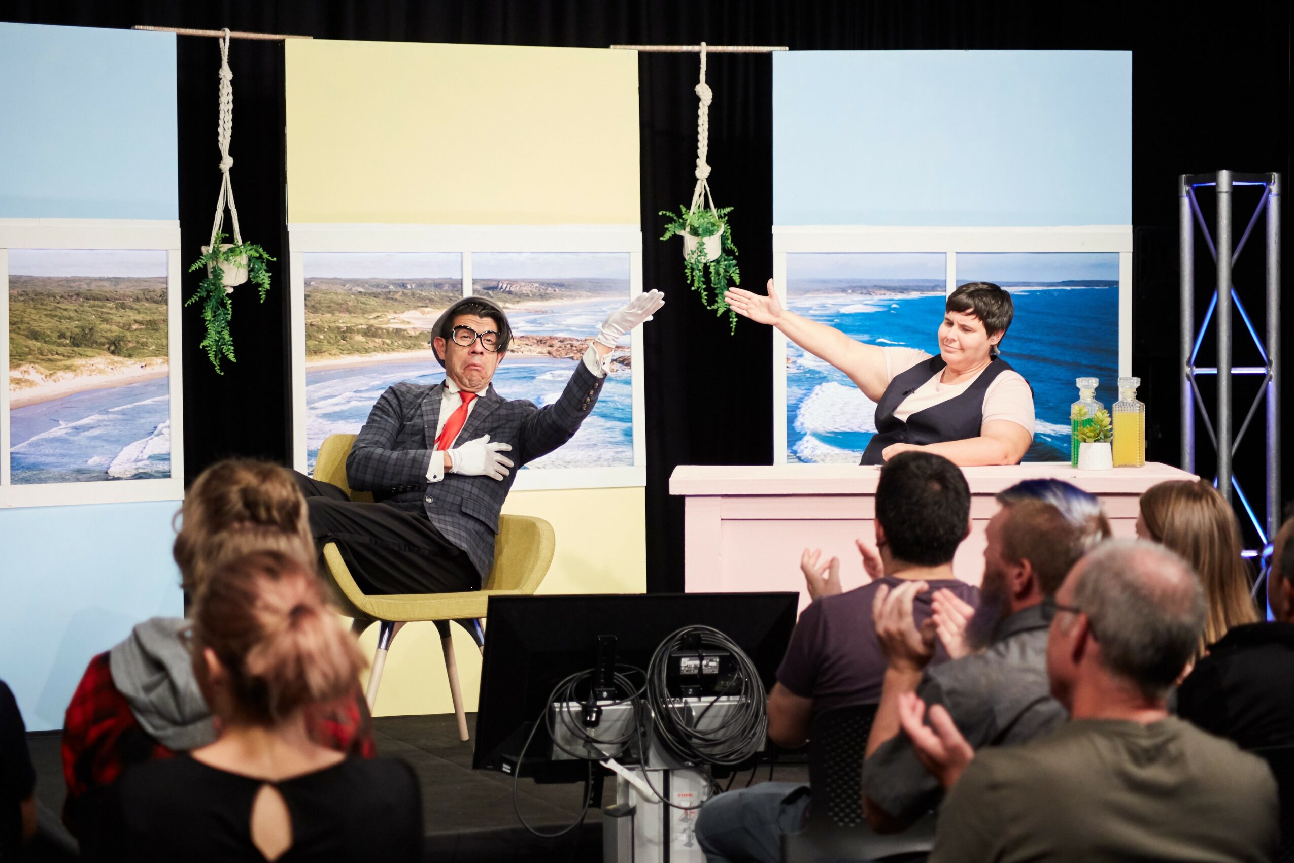 Two people on a stage with a seaside backdrop, one gesturing and the other sitting at a desk, audience watching.
