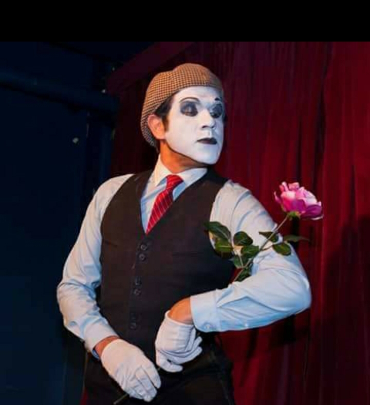 Person dressed as mime with makeup, wearing a hat, vest, and tie, holding a pink rose against a red curtain background.