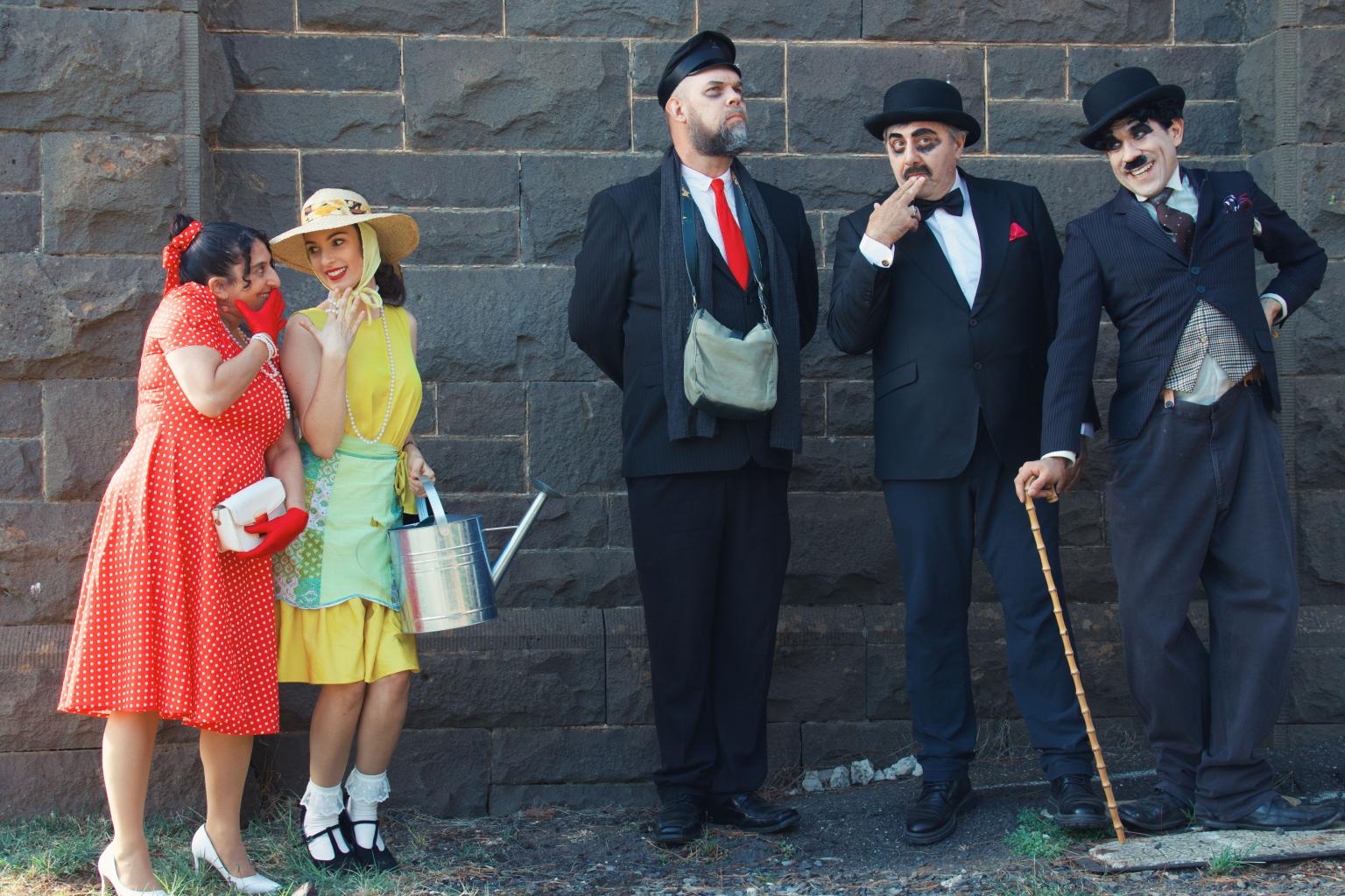 Four people stand against a stone wall, two women on the left and two men on the right, dressed in vintage clothing.