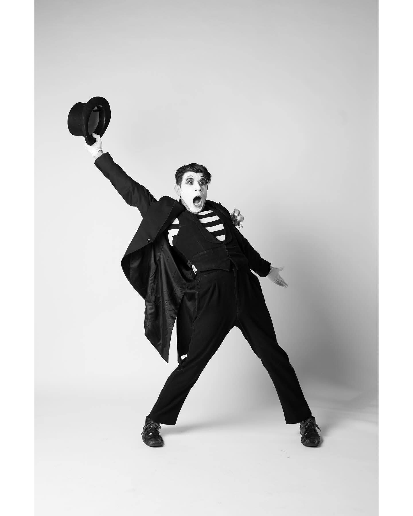 Person in a suit holding a hat, standing with legs apart and mouth open, against a plain background.