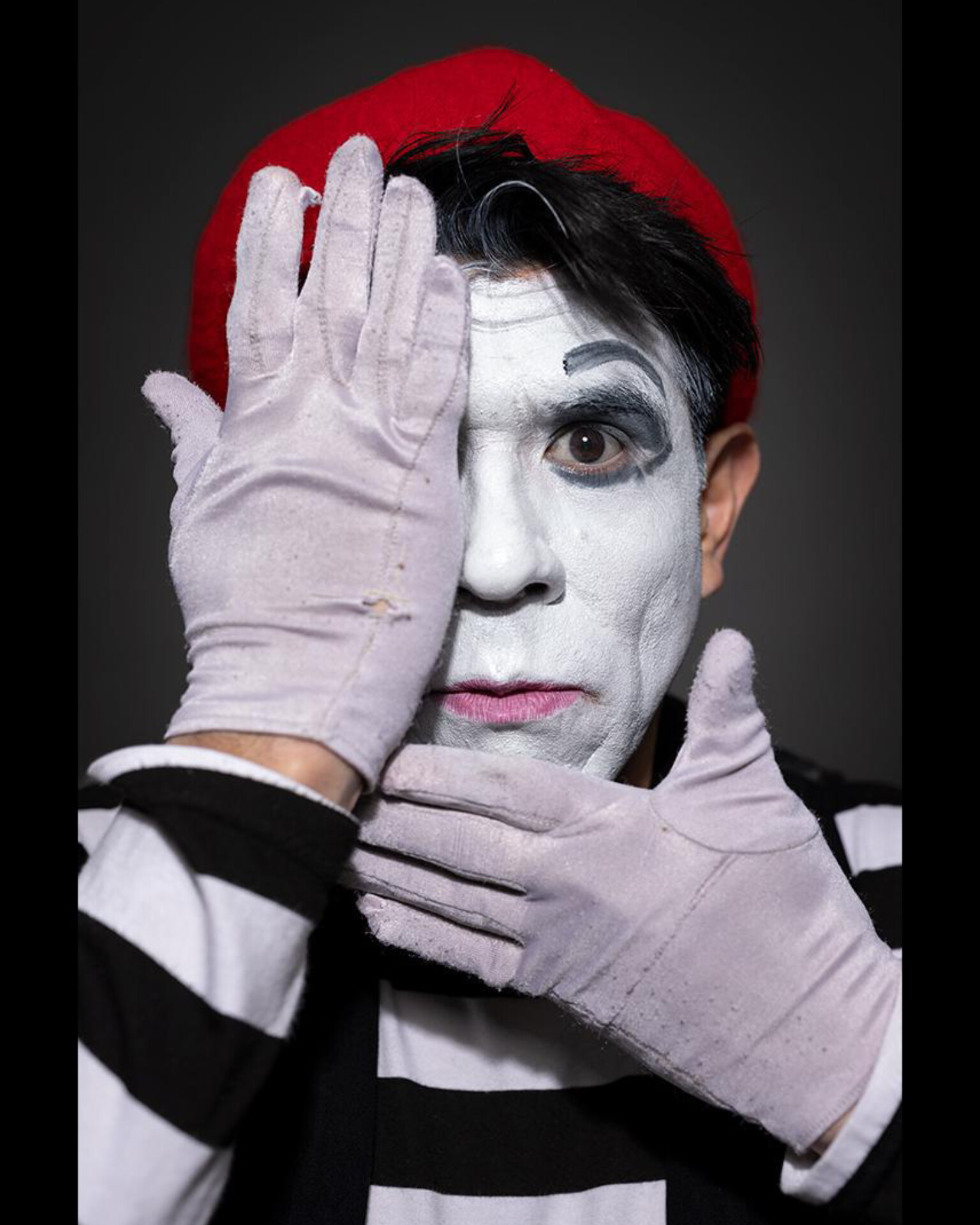Person with face painted white, wearing a red hat, striped shirt, and white gloves, covering one eye with hand.