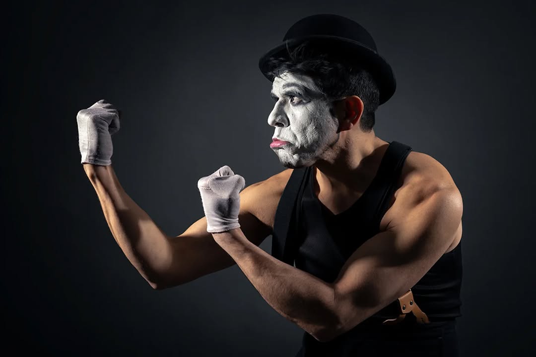 Person dressed as a mime with face paint, wearing a black hat and sleeveless top, flexing muscles against dark background.