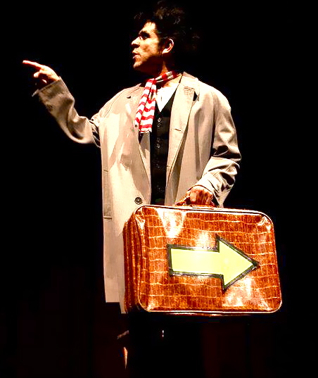 Person holding a suitcase with a large arrow sign, gesturing with their right hand, against a dark background.