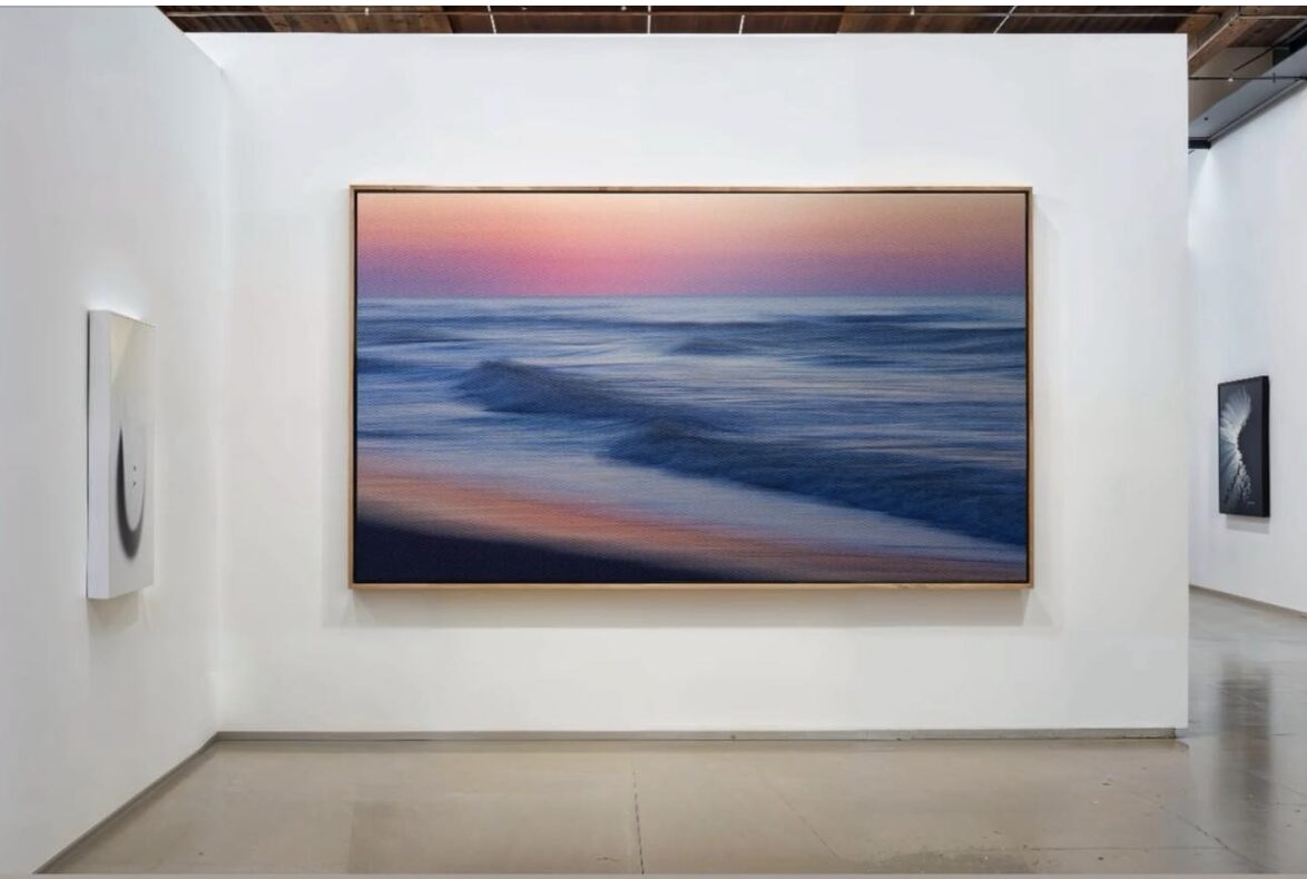Large painting of a beach scene with waves and a pink sky, displayed on a white gallery wall.