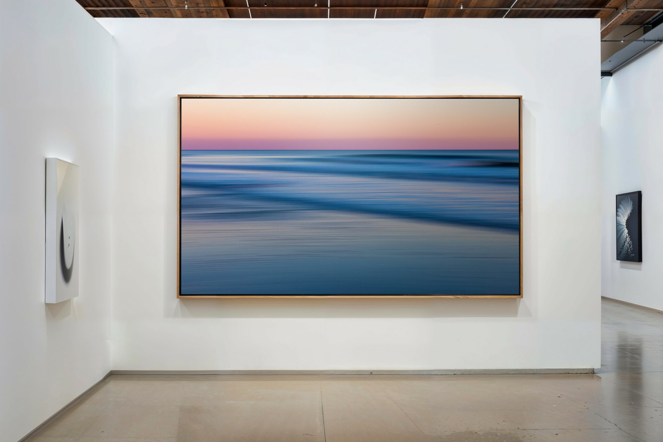 Large framed abstract painting of a seascape with pink, blue, and purple hues on a white gallery wall.