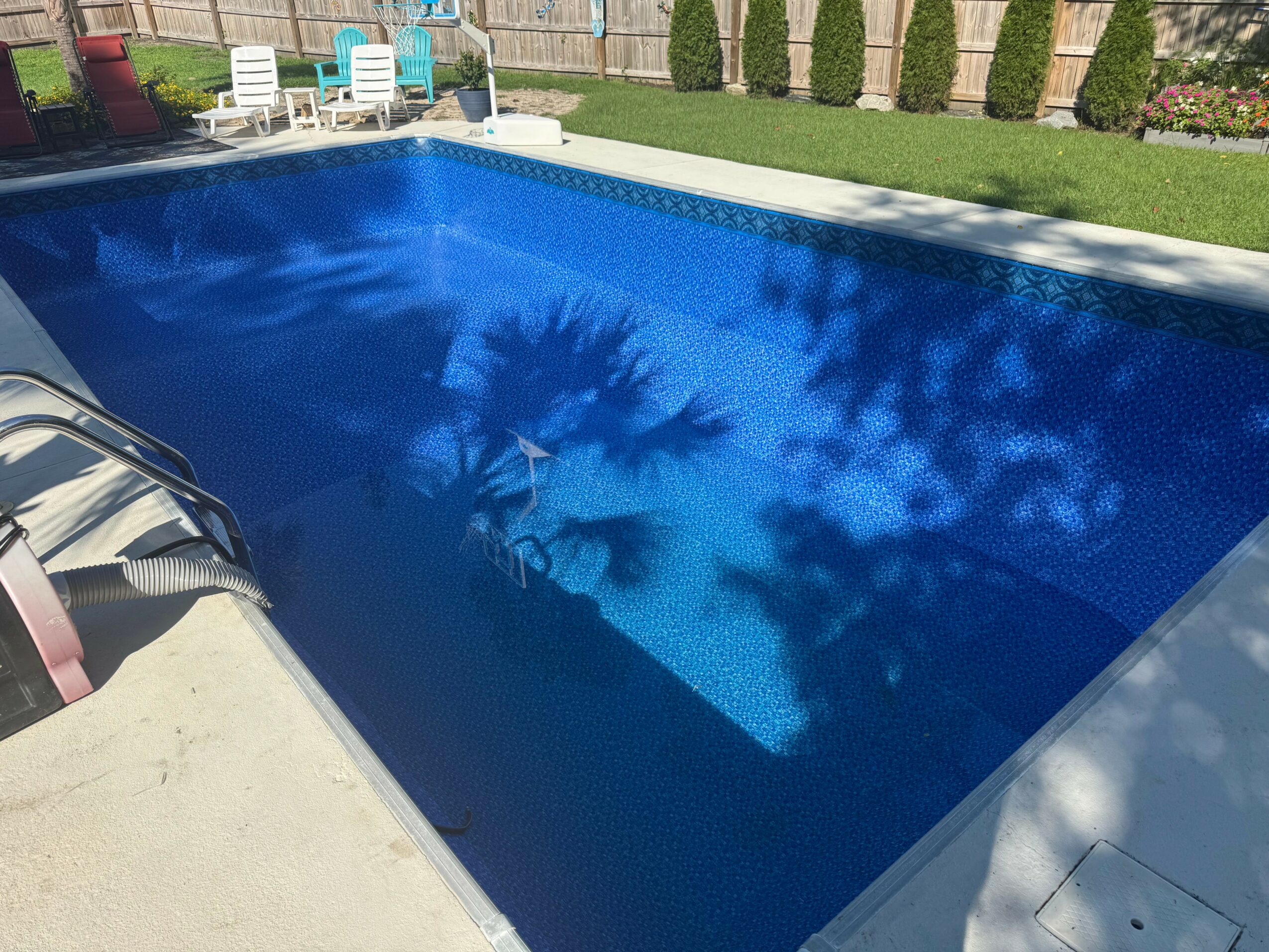Swimming pool with blue water, surrounded by a grassy area and white chairs, with shadows of trees on the water.