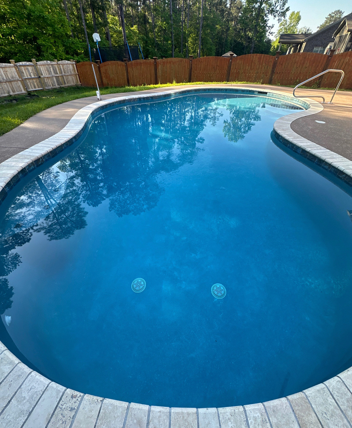 Swimming pool with clear blue water, surrounded by a deck and fenced yard, trees in the background.