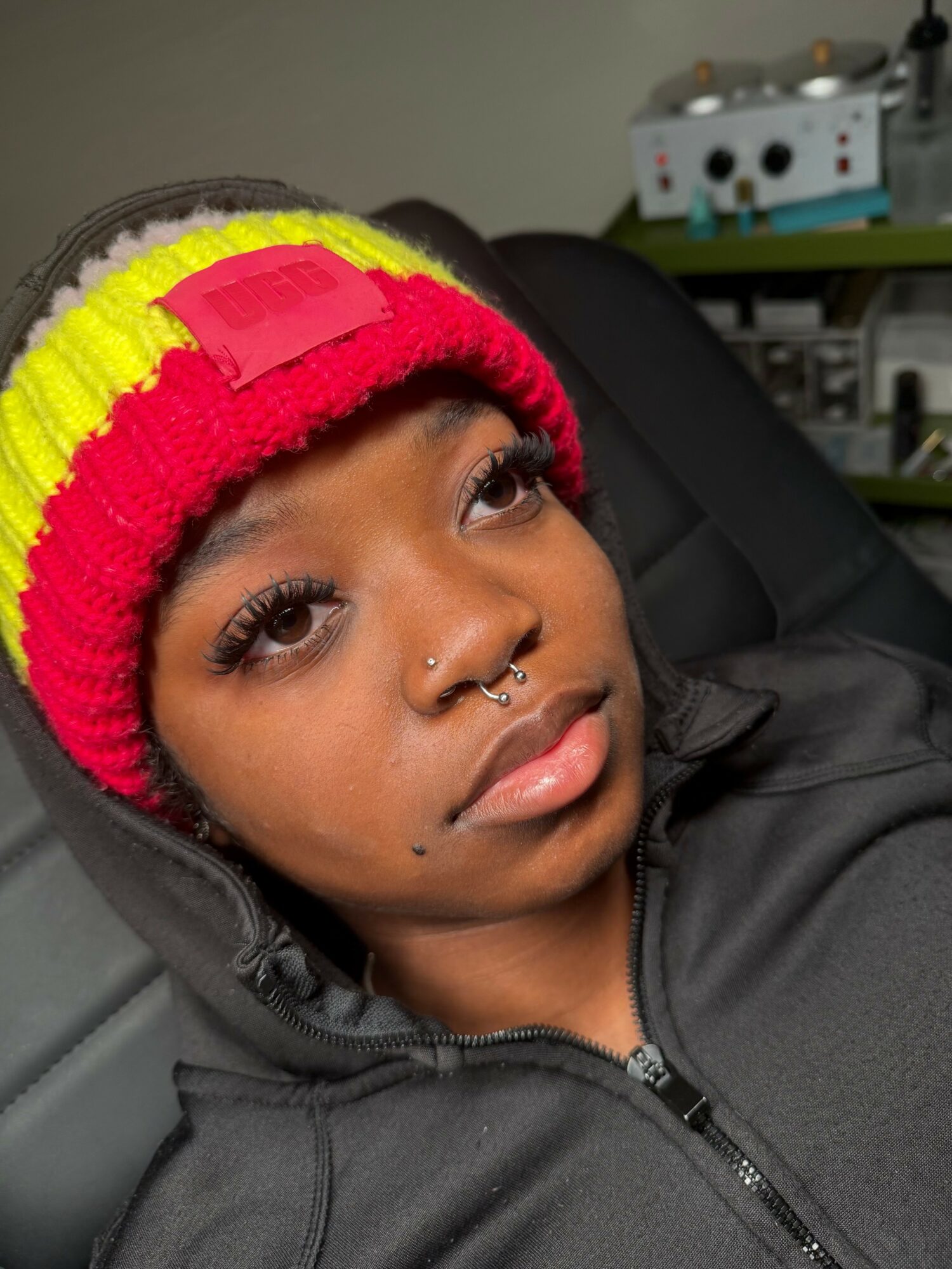 Young woman with a colorful knit hat, septum piercing, and long eyelashes, sitting in a chair indoors.