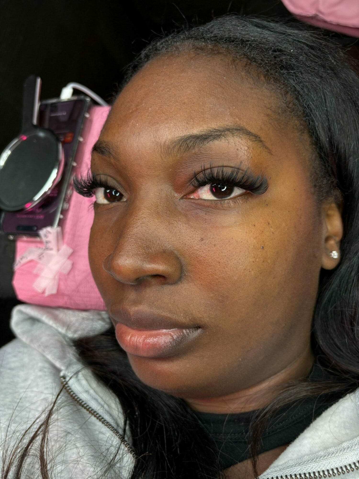 Close-up of a woman with long dark hair, makeup, and earrings, lying sideways on a pink surface with headphones nearby.