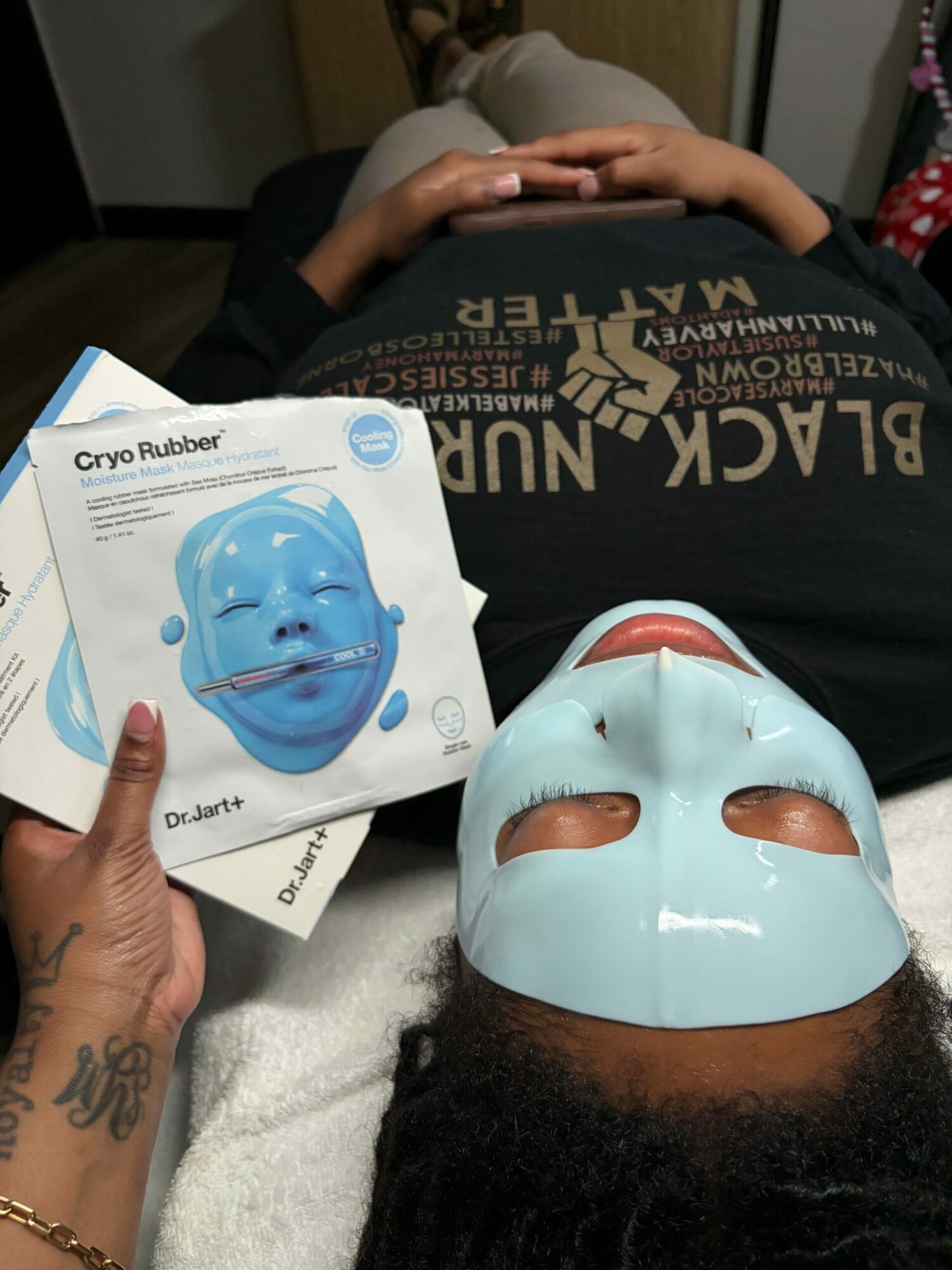 Child lying down with a light blue facial mask, holding a box of a similar mask, eyes closed, in a relaxed setting.