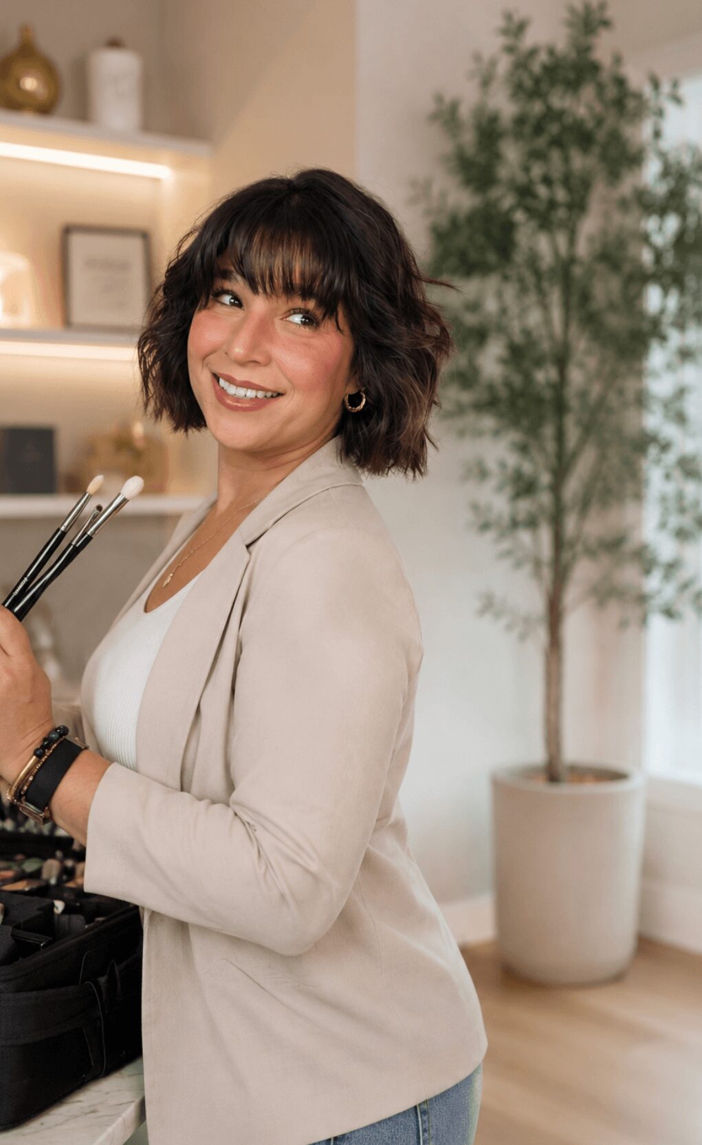 Woman with shoulder-length dark hair smiling in a modern indoor space with a potted plant in the background.