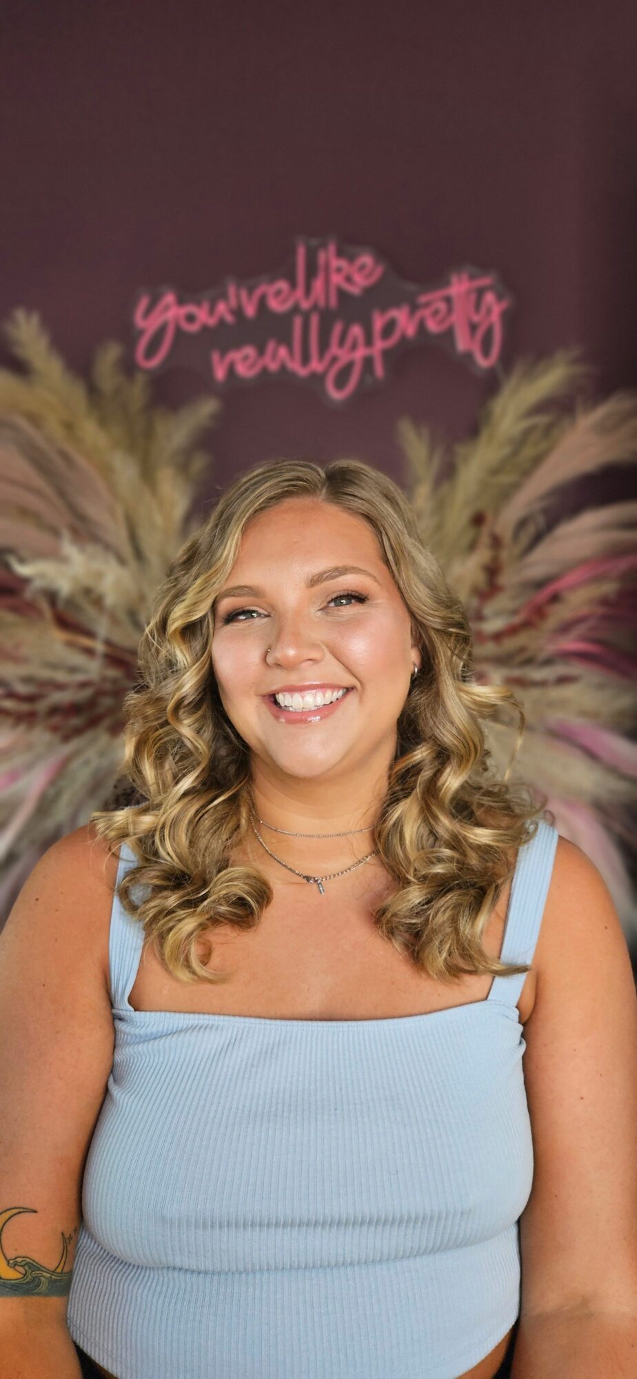 Smiling woman with curly blonde hair in a light blue top, standing in front of a dark background with pink and white text.