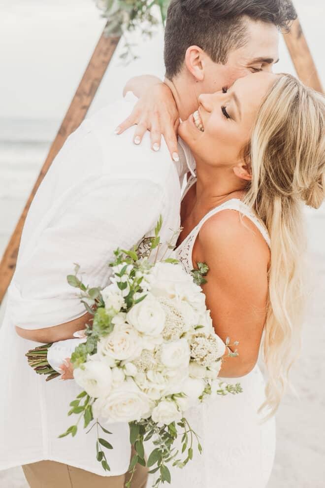 A couple embraces outdoors, holding a bouquet of white flowers, with a wooden structure and beach in background.