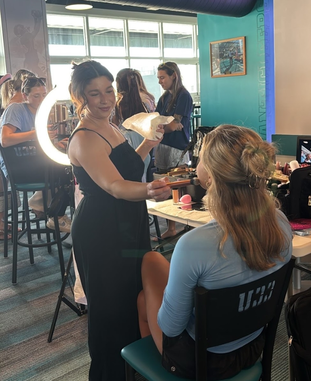 A woman in a black dress is smiling and holding a plate, talking to a seated woman with long hair in a blue top, in a room with large windows and other people. There is a ring light in the background.