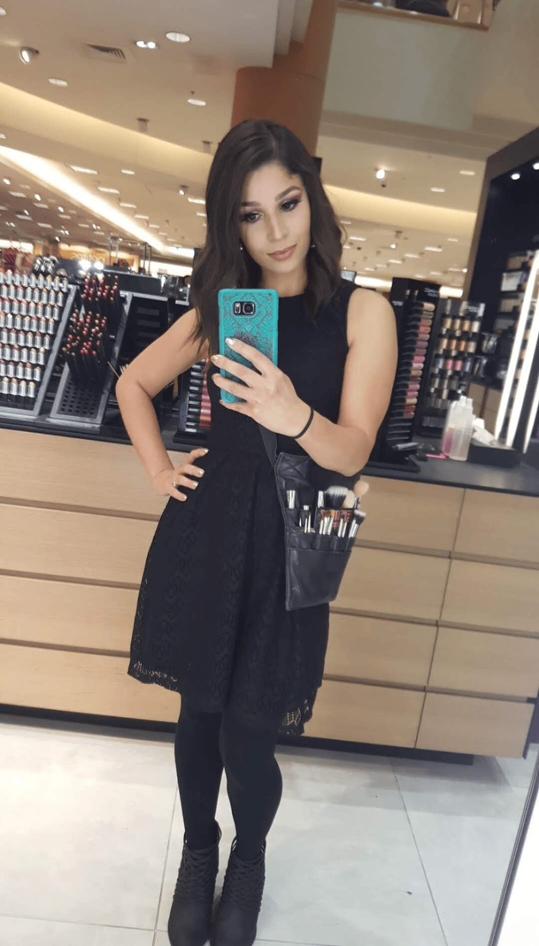 Young woman with long dark hair taking a mirror selfie in a store, wearing a black dress and boots, holding a shopping bag.