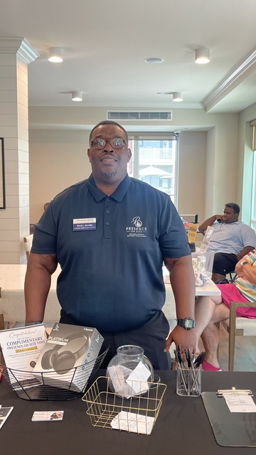 Man in navy polo shirt standing behind a table with promotional materials, in a bright indoor setting.