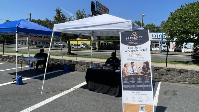 Outdoor event with tents, a table, and a signboard in a parking lot, with trees and buildings in background.