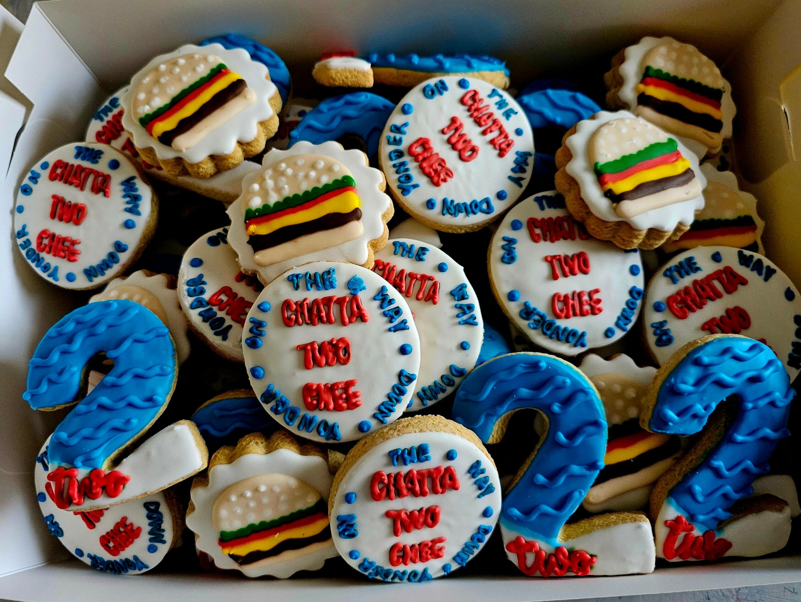 Box of cookies decorated with rainbow stripes, blue number 2, and text celebrating a birthday, with colorful icing and designs.