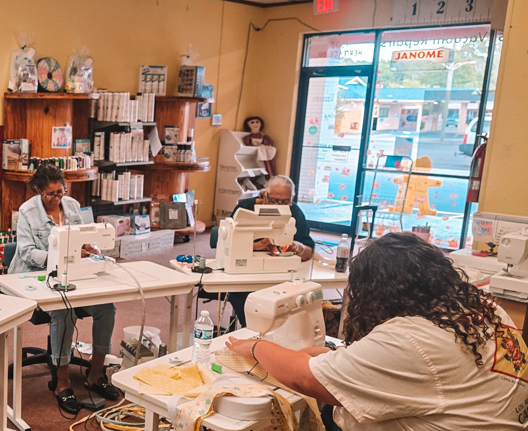 People working at tables with sewing machines inside a store, with a large window showing street view outside.