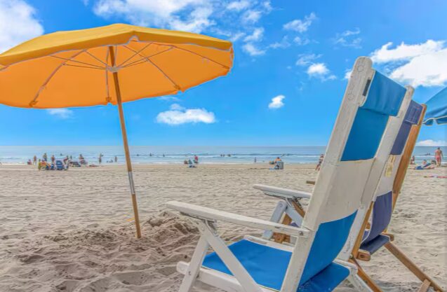 Beach scene with a yellow umbrella and two white and blue chairs on sand, ocean and sky in background.