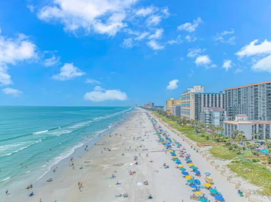 Beach with umbrellas, buildings, and a blue sky with clouds, viewed from above.