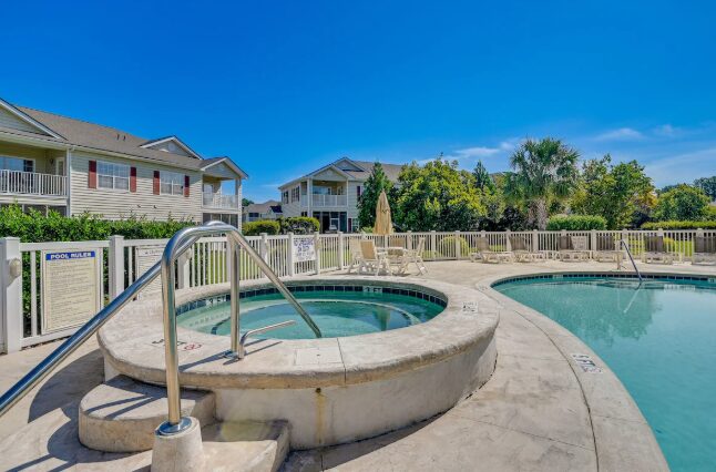 Swimming pool with hot tub, surrounded by a fence, with houses and trees in the background under a clear blue sky.