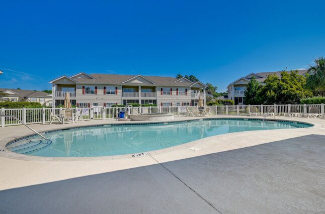 Outdoor swimming pool with a fence, surrounded by apartment buildings and trees, under a clear blue sky.
