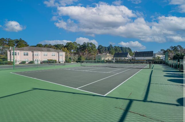 Tennis court with green surface, white lines, surrounded by a fence, under a blue sky with clouds.