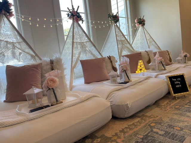 Four beds with pink pillows and teepee tents decorated with flowers and string lights, set up in a room with large windows.