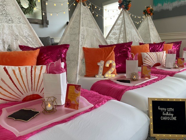 Decorated beds with pink, orange, and purple pillows, tents, and table settings for a celebration, with string lights overhead.