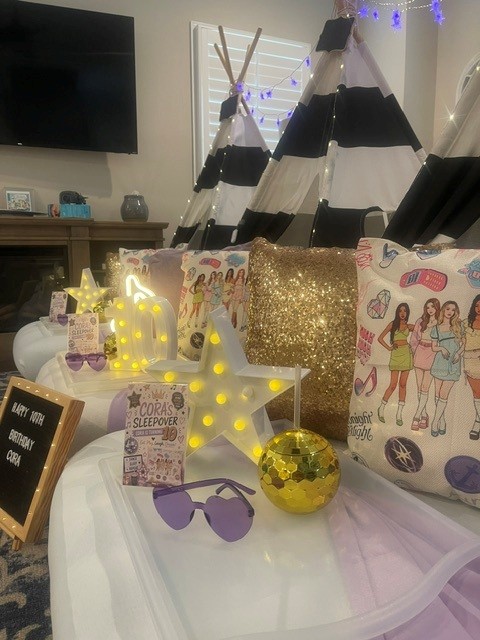 Party decorations including a star-shaped light, sunglasses, a gold glitter box, and a bag with girls in swimsuits, on a table.